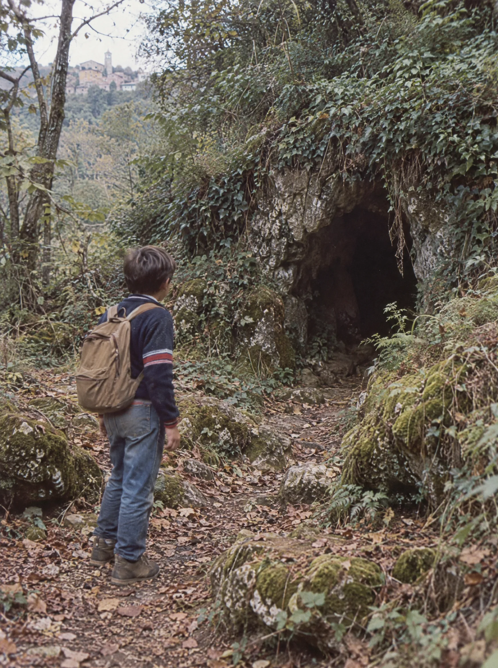 Scena fotografica ricreata: un ragazzo di spalle davanti a una grotta nel bosco